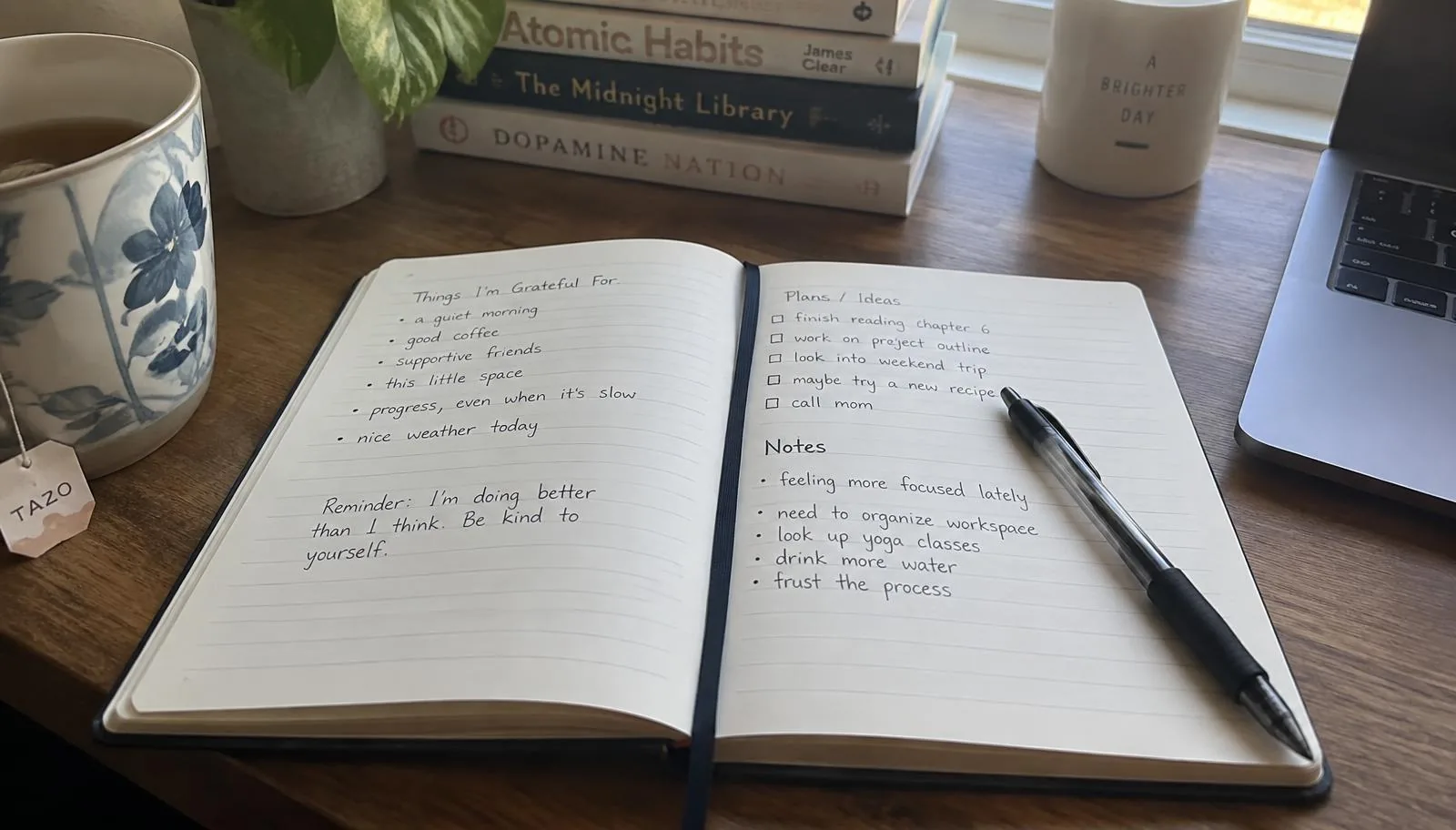 An open journal with handwritten gratitude lists beside a pen, tea, and a stack of books