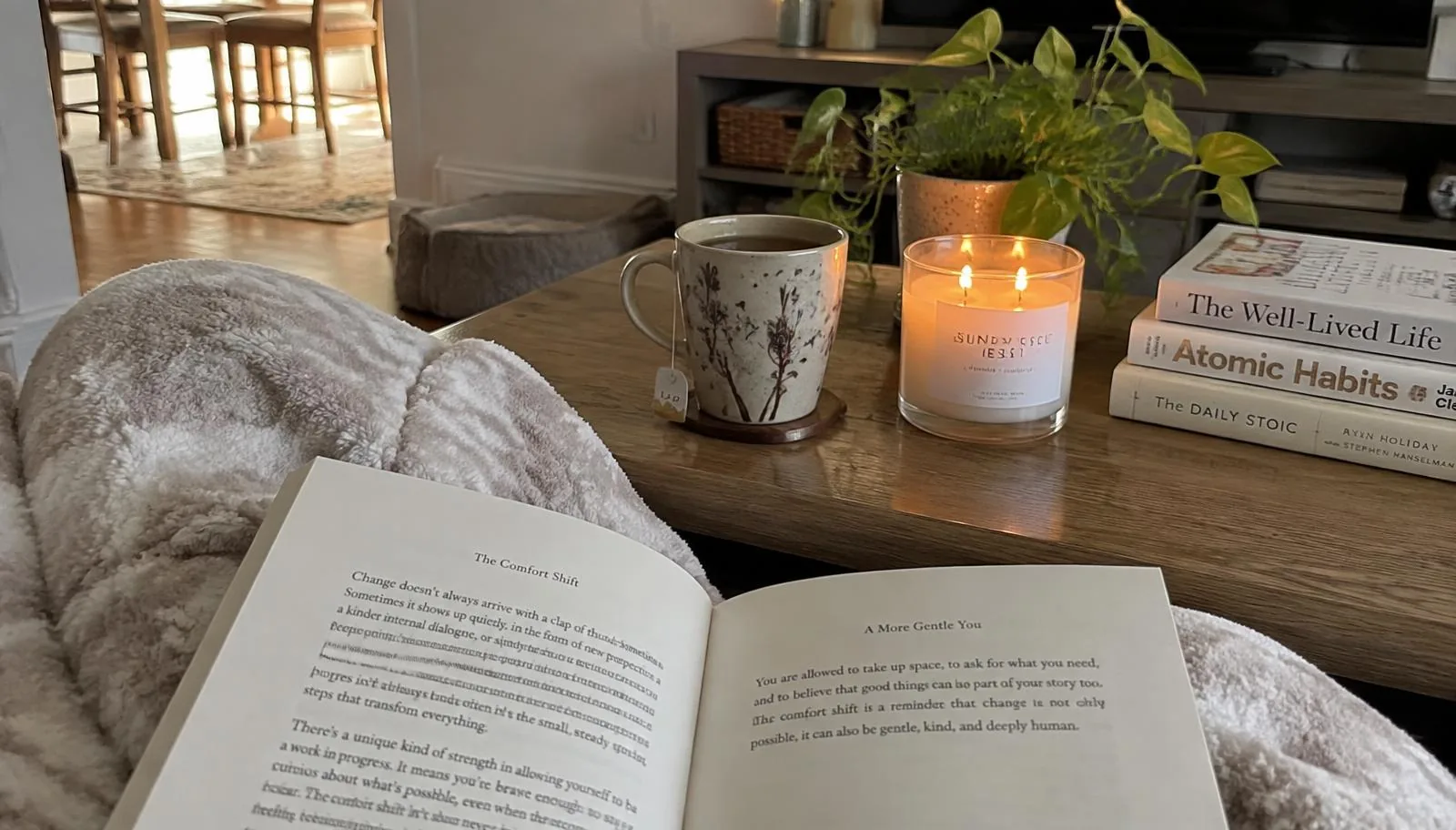 A cozy scene with a soft blanket, an open book, a mug of tea, and a lit candle on a coffee table